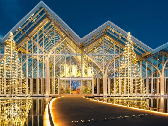 A glass conservatory with peaked roofs, seemingly floating on water, lit up in white.
