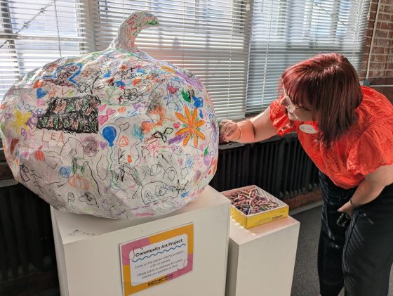 A person with shoulder length red hair bending over to sign a paper-mache pumpkin.