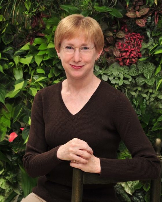 A head shot of a person in a brown shirt, with short blond hair, and gold glasses, smiling at the camera.