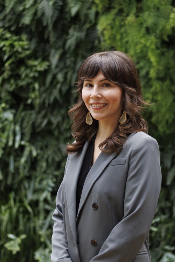 A white woman with long brown hair wearing a grey suit stands in front of a leaf-covered wall. 