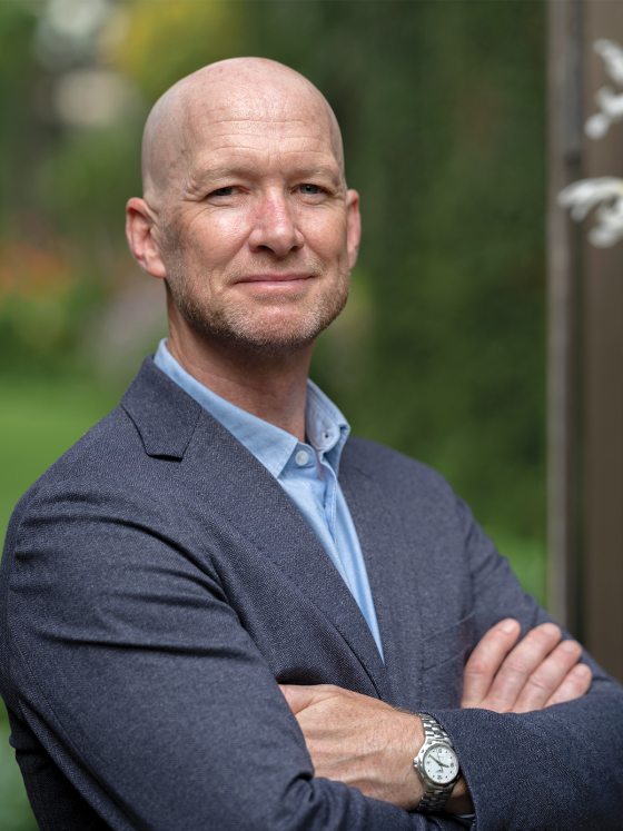 Headshot of bald man wearing blue suit jacket with arms crossed