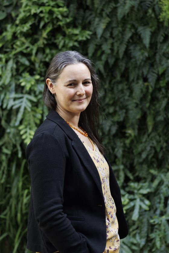 A white woman with grey hair wearing a black cardigan and yellow shirt printed with flowers stands in front of a leaf-covered wall.