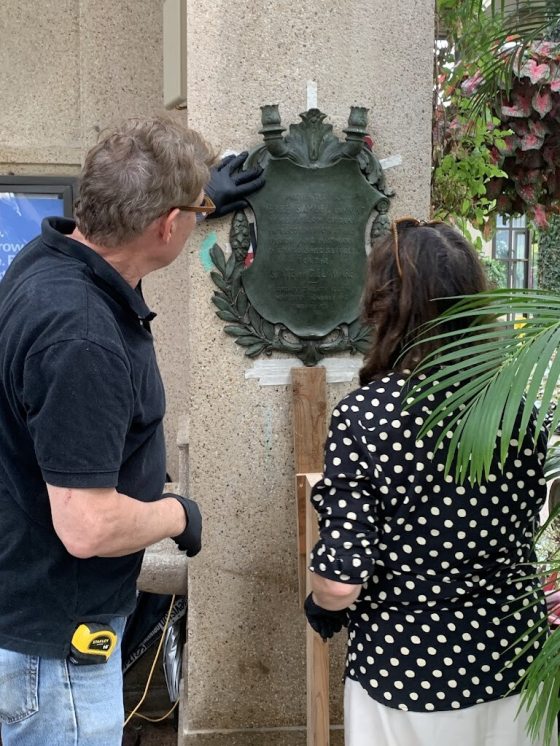 Two people examining a bronze plaque hanging on a column in a conservatory.