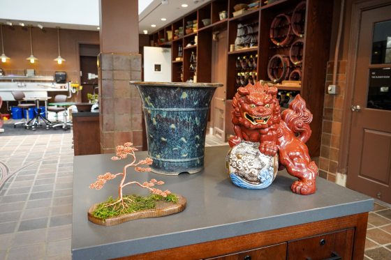 A foo dog, antique Chinese pot, and wire bonsai all on a table inside a bonsai workshop.