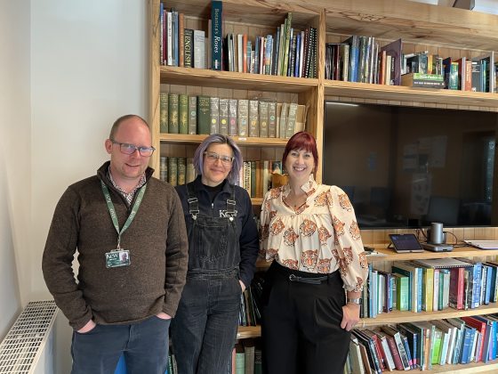 Three people standing shoulder to shoulder smiling at the camera, in front of a large bookshelf.
