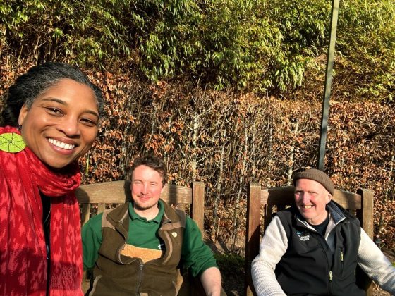 Two people sitting and one person standing, taking a selfie outdoors.