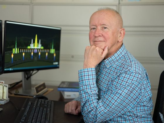 A person in a blue and gray plaid shirt, sitting at a control booth, resting their head on their hand and looking at the camera.