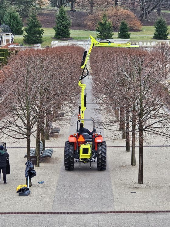 A red tractor with a tall yellow shearing device that is being used to practice trimming trees in winter.