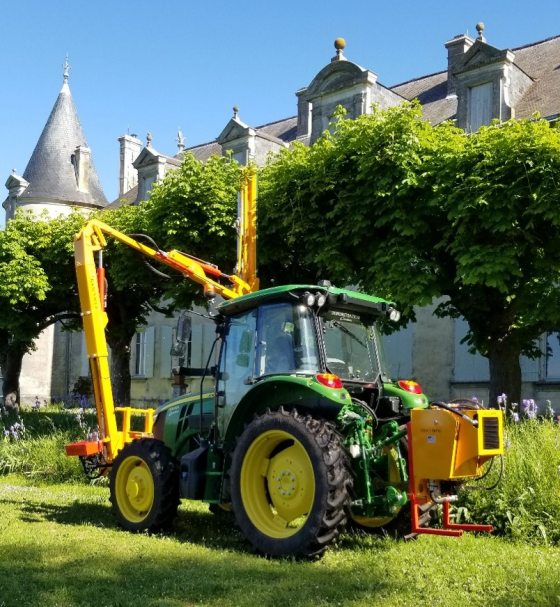 A green tractor with a tall yellow shearing device that is being used to trim trees.