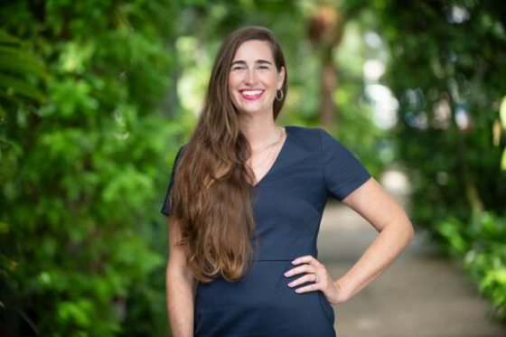 A woman with brown hair wearing a navy dress smiles for the camera while being surrounded by greenery. 