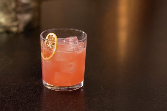 A salmon colored cocktail, in a highball glass, garished with dried orange, on a black countertop.