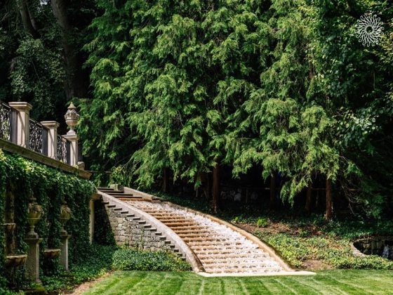 A curved stone staircase and stone facade in an outdoor garden, with tall evergreens in the background.