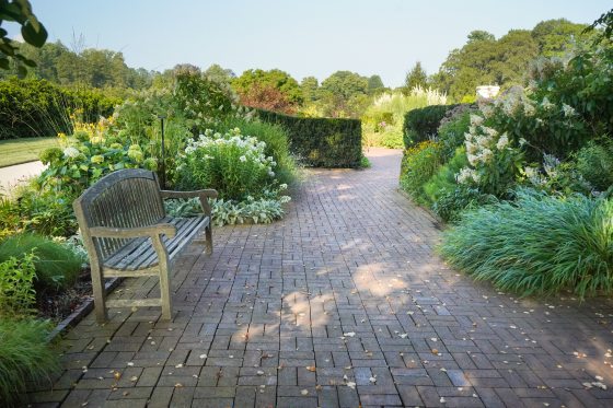 A wooden bench, set along a brick path, surrounded by lush summer foliage.