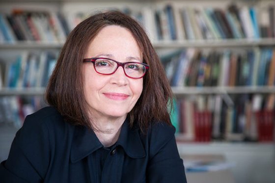 A head shot of a person with shoulder length brown hair, red glasses, and a black shirt smiling at the camera.
