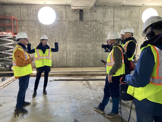 A group of people in white hard hats and yellow saftey vests, filming in a concrete tunnel.