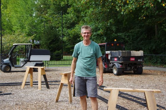 A person outdoors on a gravel ground. In the background, there is a golf cart on the left and a small, red utility vehicle with a dump bed on the right. Several wooden sawhorses or simple trestle structures are scattered on the ground near him, with some model train tracks visible in the very bottom foreground.