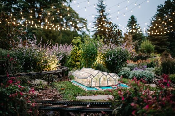 An outdoor, dusk-time view of a lush garden display featuring a model railway. A miniature, illuminated greenhouse with an arched roof and a small blue pond sits in the center of the frame. Model train tracks curve around the greenhouse.