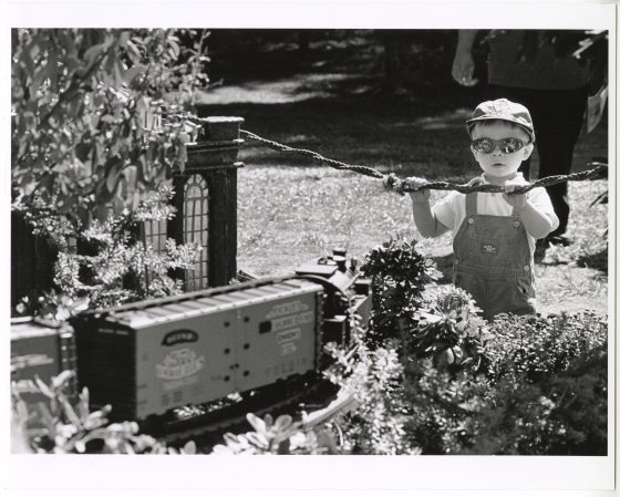 A black and white photograph of a young child, wearing sunglasses, a cap, and denim overalls, holding onto a rope barrier while observing a large-scale model train set.