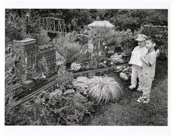 A black and white photograph of two young children, standing on a patch of grass and smiling as they look at a large, elaborate garden model railway setup. 
