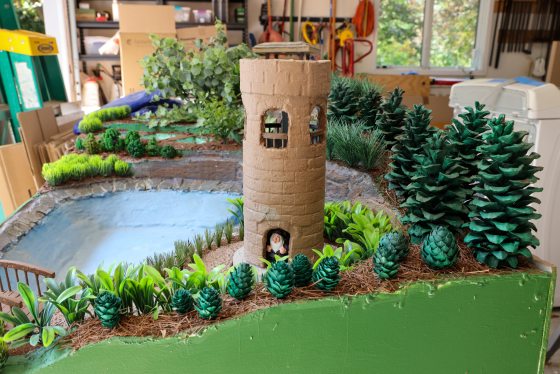 A detailed, close-up view of a miniature garden landscape model, built on a green platform. The scene features a tall, cylindrical, light-brown tower with arched openings, standing near a small, blue-colored pond. 