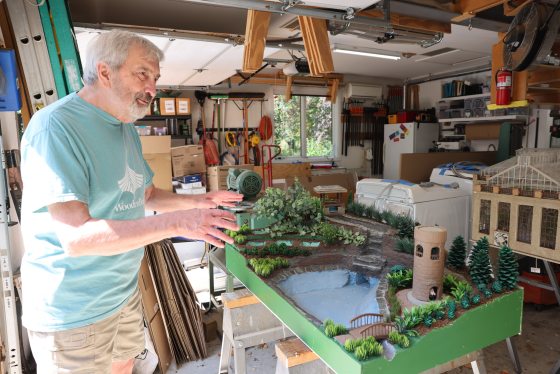 A person with gray hair and a light blue t-shirt stands in a garage or workshop, enthusiastically gesturing with both hands toward a detailed, elevated miniature landscape model. The model features a blue pond, a bridge, a round brick tower, small evergreen trees, and lush green foliage.