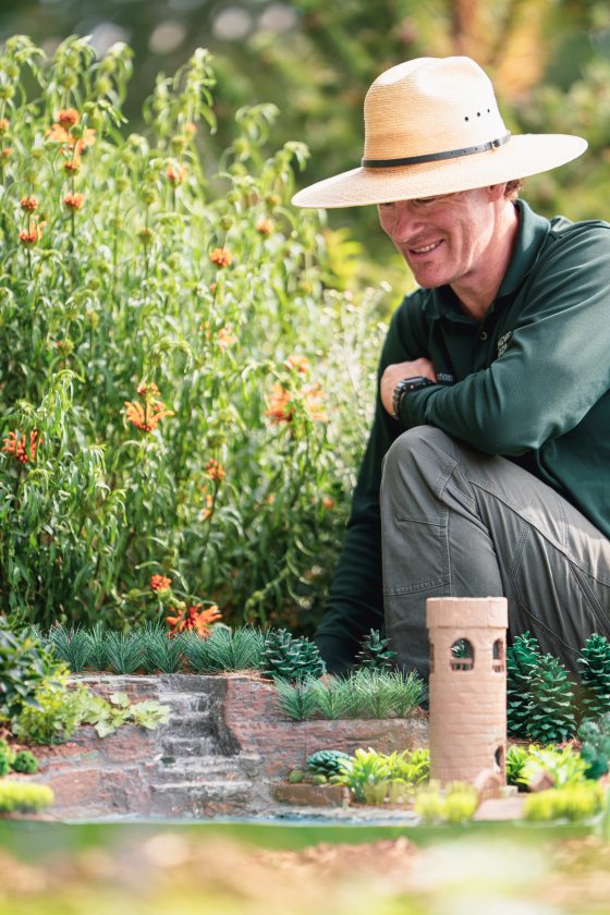 A vertical shot showing a man in a wide-brimmed straw hat and dark green shirt crouched down, smiling as he tends to or observes an outdoor miniature garden and model railway scene.