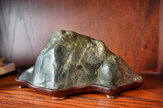 A close-up, dimly-lit photo of a smooth, dark green and black mottled stone, shaped like a low, rolling hill or mountain. It is set into a custom-fitted, dark brown ceramic or wooden tray with short, rounded feet. The object is resting on a dark wooden shelf, with a warm, reddish-brown wooden paneling in the blurred background.