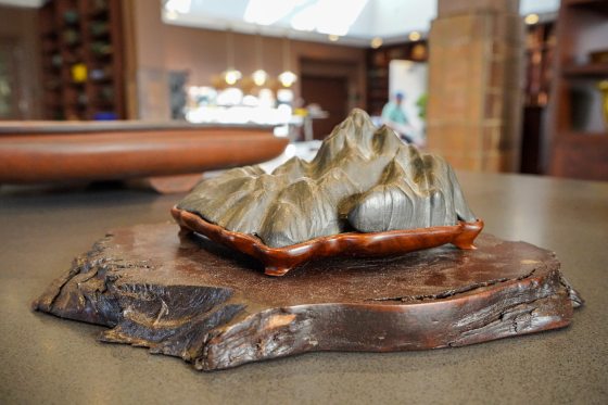 A low-angle, close-up shot of a small, dark grey rock carving, shaped like a miniature mountain range with multiple jagged peaks. This carving is set into a custom-fitted, dark reddish-brown wooden tray with curved feet. The entire piece rests on a larger, thick, natural-edge slab of dark, rustic wood that is sitting on a grey table. The background is a brightly-lit, blurred indoor space with brown and tan tones.