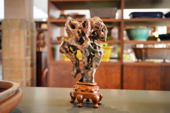 A close-up, front-facing shot of a slender, vertical, highly-eroded, dark reddish-brown rock or wood carving with multiple openings, resembling a stylized, abstract mountain peak or column. It is mounted on a carved, highly-polished, dark wooden stand with four bulbous, claw-like feet. The object sits on a grey table with a blurred background of wooden shelves displaying pottery and other objects.