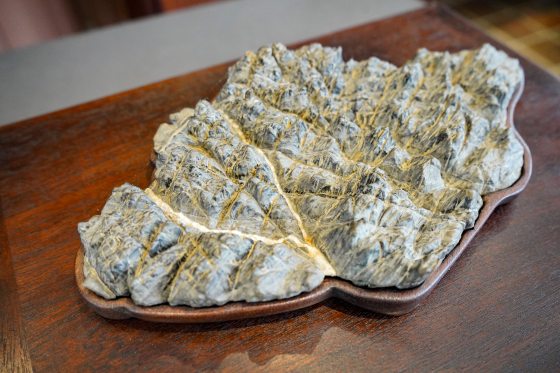 A close-up, high-angle shot of a flat, relief carving made from a grey-blue, highly-fissured stone, textured to resemble a miniature, snow-capped mountain range or topographical map. It has a prominent diagonal white seam running through it. The stone is set into a custom-fitted, shallow, reddish-brown tray or base. The object sits on a dark brown wooden surface, with a grey background blurred behind it.