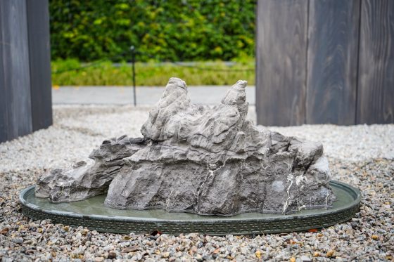A low-angle, eye-level shot of a large, grey, craggy rock formation with stratified layers, resembling a low mountain or island. The rock is sitting in a shallow, circular, dark grey metal tray that contains a small amount of water, creating a miniature landscape. The tray rests on a bed of light grey and tan gravel. In the background, there are two dark wooden vertical posts framing a bright, blurred view of green foliage.