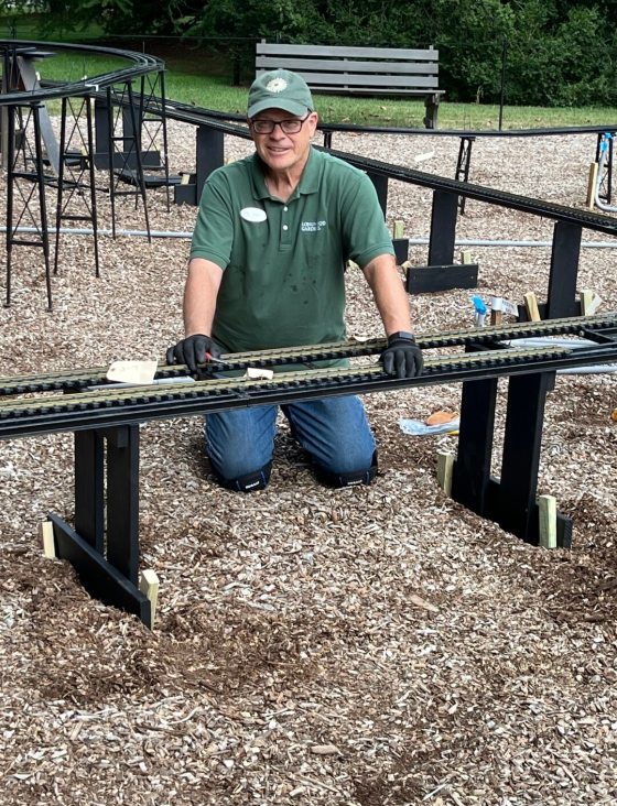 A vertical shot of a person kneeling outdoors, working on a raised section of black model train track set on trestle supports over a wood-chip ground. He is wearing a green polo shirt, jeans with kneepads, black gloves, and a green cap, and is looking directly at the camera. 