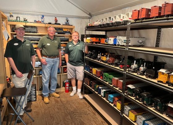 Three people wearing matching green polo shirts stand together, smiling, inside a shed that serves as a display room for model trains.