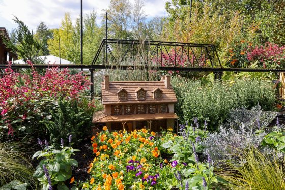 An outdoor, eye-level view of a miniature garden display featuring a detailed model of a small, rustic wooden house with three dormers. The house is surrounded by a lush and colorful garden with clusters of vibrant orange, purple, and magenta flowers and green and silvery foliage. 