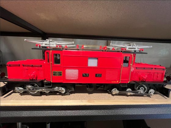 The model is painted a bright red with black and silver details, and it features silver pantographs on the roof. It is displayed on a tan surface on a black metal shelf.