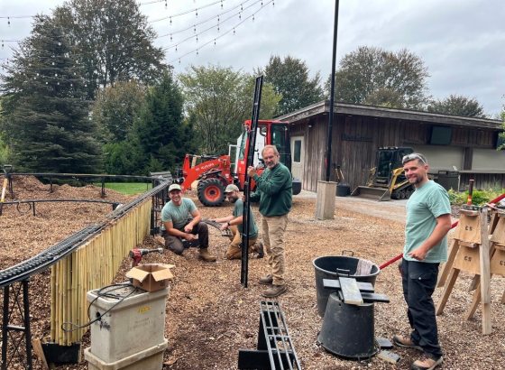 A group of four people working on setting up infrastructure for a model railway. Three are gathered around a newly installed black pole, possibly for string lights, with one is standing in the foreground, smiling. The ground is covered in wood chips, and in the background, there is a partially finished model train track alongside a wooden fence, construction equipment (a red tractor), and a wooden structure.