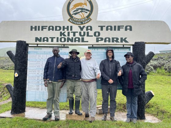 A group of five people are standing in front of a large wooden sign for Kitulo National Park in Tanzania. They are all smiling, with three of them giving a "thumbs-up" gesture. The sign is written in both Swahili and English.