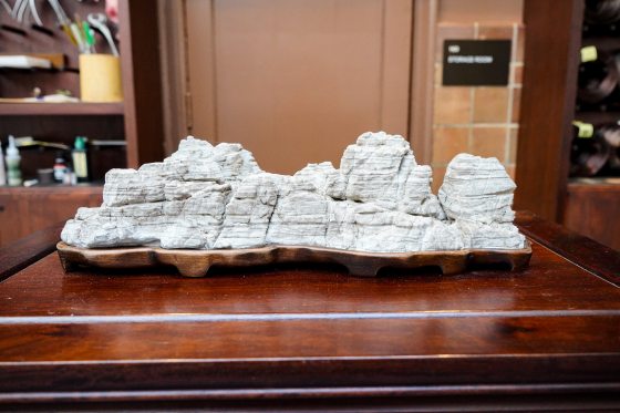 A low-angle, eye-level shot of a large, elongated Suiseki (viewing stone) or Penjing accessory, carved from a light grey or white stone to resemble a craggy, stratified mountain range. It is mounted on a custom-fitted, dark brown wooden stand with several low feet. The entire piece rests on a highly-polished, dark wooden table or cabinet, with a blurred background of warm brown paneling and a dark sign.