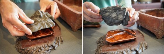 A close-up shot of a person's hands holding a dark grey, smooth, and highly fissured rock, shaped like a miniature mountain or undulating peak, suspended over a table. Below the rock is a small, custom-fitted, highly-polished, reddish-brown wooden tray. Both the rock and tray are positioned over a larger, thick, natural-edge slab of dark wood, which sits on a grey table. A rustic, rectangular, reddish-brown ceramic pot is visible to the right.