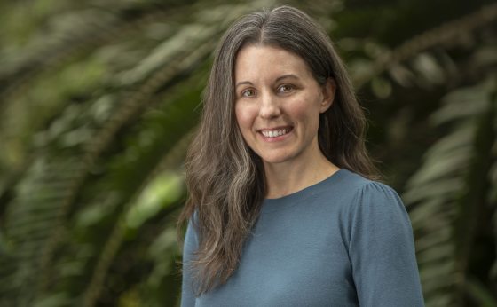 An outdoor, close-up photograph of a woman with long, wavy brown hair, wearing a teal or dusty blue long-sleeved top. She is smiling directly at the camera. She is positioned against a soft-focus, deep green background of dense foliage and large ferns.