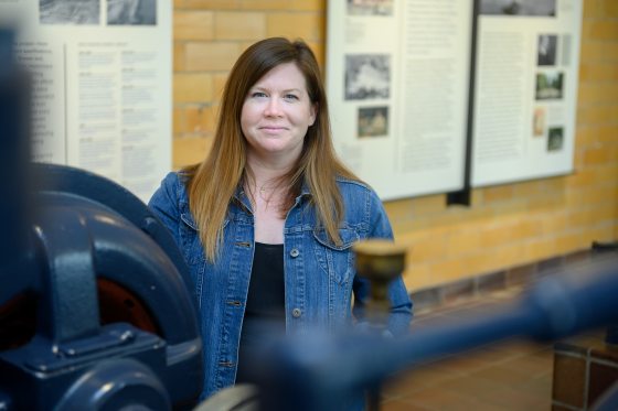 A smiling woman with long brown hair, wearing a denim jacket, stands inside a museum exhibit next to large, blue industrial machinery and information display boards.