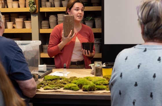An instructor in an orange cardigan holds up a rectangular piece of wood or brick to a small audience during a terrarium or moss gardening workshop.