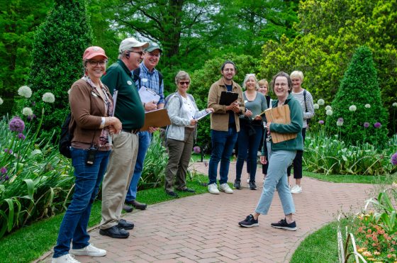 A group of seven adults, some holding clipboards, stands on a brick pathway in a lush garden, smiling toward the camera during an outdoor tour or class.