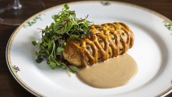 A golden-brown individual Beef Wellington or pastry en croute, with an intricately woven lattice crust, plated on a white dish with a pool of creamy, light brown sauce and a generous mound of fresh micro greens on top.