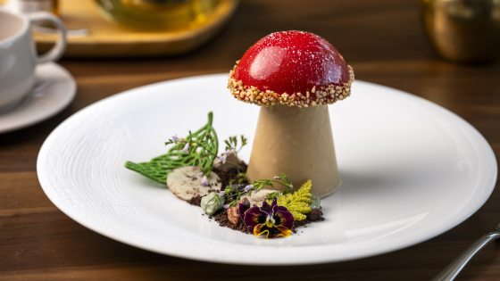 A dessert shaped like a large, stylized toadstool or mushroom, with a glossy red, nut-crusted cap and a light brown stem, plated on a white dish with surrounding garnishes including micro greens and an edible pansy flower.