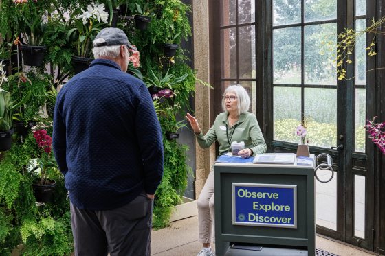 A staff member engages in conversation with a man, whose back is to the camera, at a small information kiosk labeled "Observe, Explore, Discover" within a greenhouse setting.