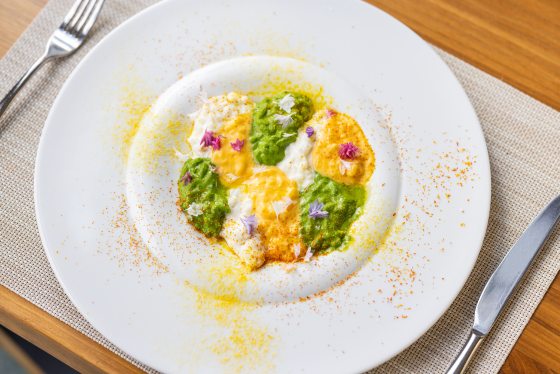 A gourmet dish, possibly a risotto, served on a white plate with fork and knife on either side, decorated with dollops of white, yellow, and green sauces, edible flowers, and colorful spice dust.