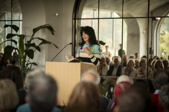 Poet Joy Harjo, wearing a striped sweater, stands at a light wood podium reading from a book to a seated audience in a large, bright room with arched glass windows overlooking a garden.