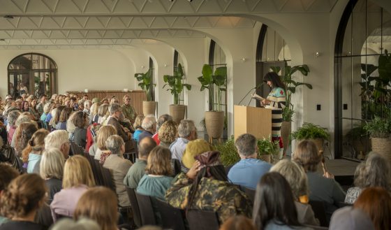 Wide shot of an indoor reading or lecture event. Poet Joy Harjo is at a podium on the right, reading to a large, seated audience filling the foreground. The room features a white arched ceiling and tall potted plants.