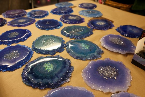 An array of shiny, finished resin geode slices in shades of blue, turquoise, and purple, with sparkling centers, laid out on a brown surface.